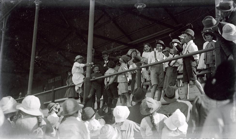 Large froup of children at Alberton Oval looking up at boys in front row of grandstand trying to eat donuts from a horizontal string. c 1920