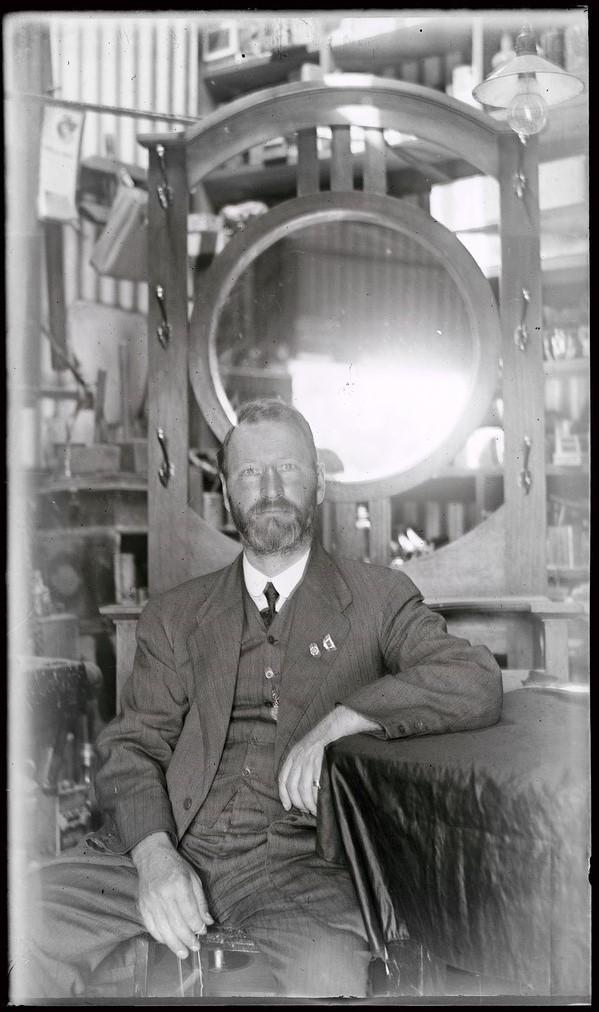 William Charles Brooker seated in front of mirrored hall, probably in warehouse of his furniture shop, c 1920