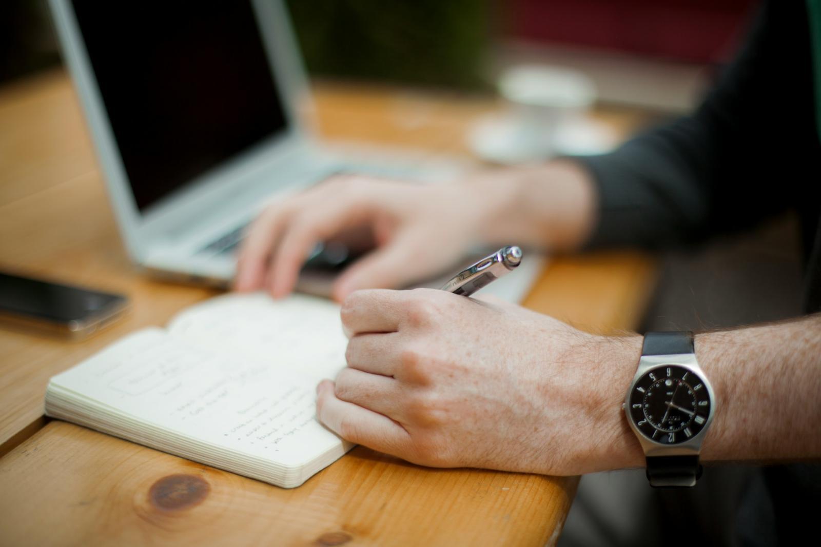 Photo of a laptop on a desk and a man writing in a notebook.