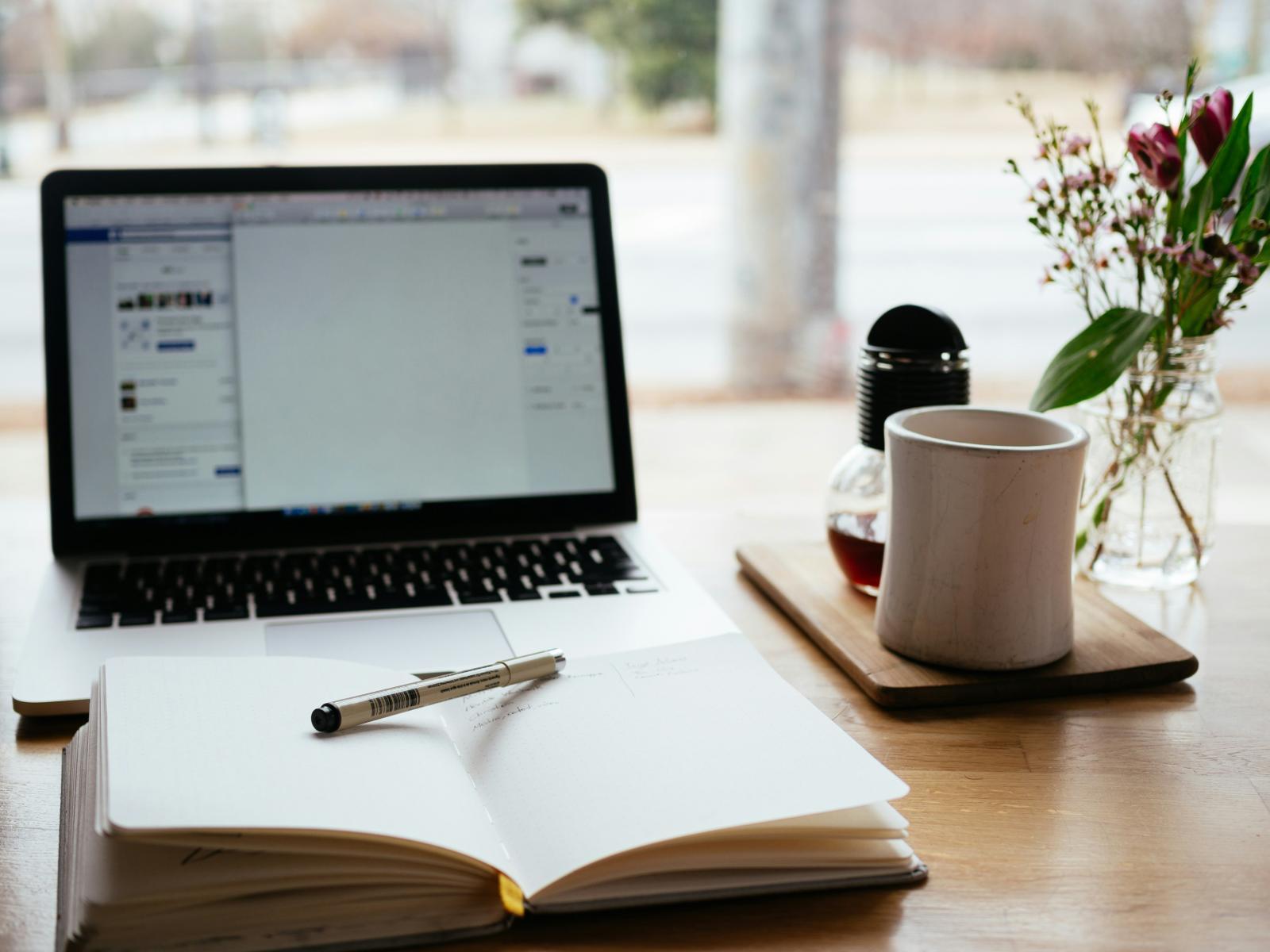 Computer on a desk with a coffee cup, note book, pen and a vase with flowers