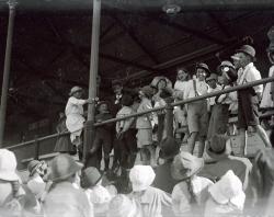 Large froup of children at Alberton Oval looking up at boys in front row of grandstand trying to eat donuts from a horizontal string. c 1920