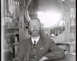 William Charles Brooker seated in front of mirrored hall, probably in warehouse of his furniture shop, c 1920