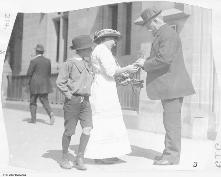 Woman selling wattle sprig to man on city street, watched by young boy, 1918